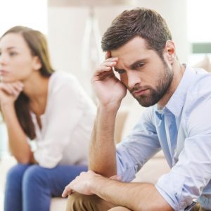 Relationship breakdown. Depressed young man holding hand on head and looking away while woman sitting behind him on the couch