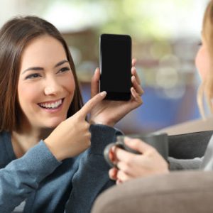 Happy girl showing a blank phone screen to a friend on a couch at home